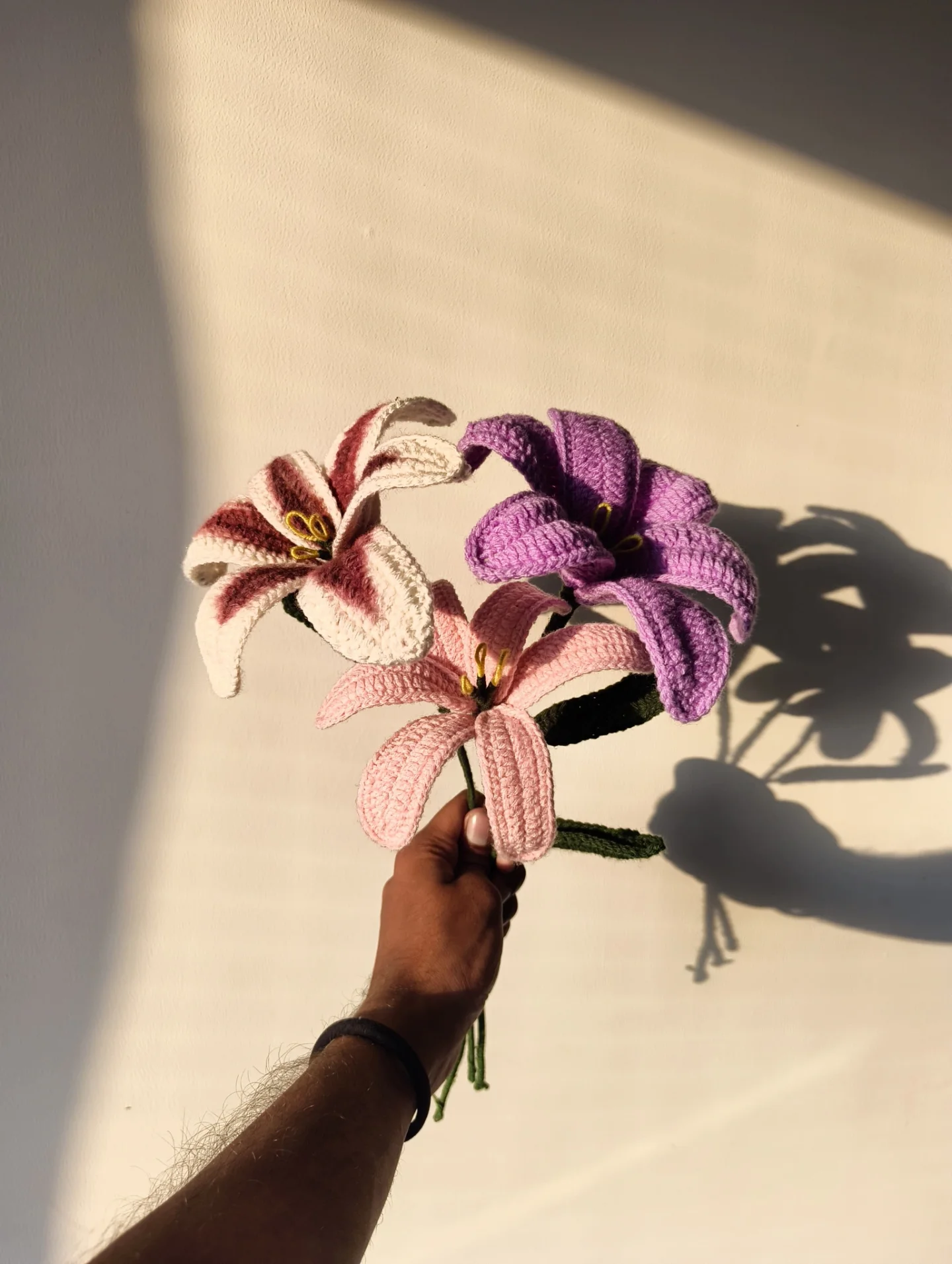 A trio of crochet lilies — pink, blush and lavender — held up against a sunlit wall.